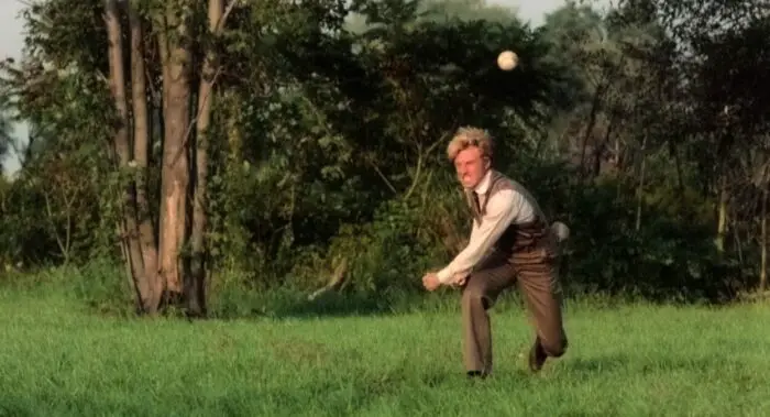 Roy Hobbs throws a baseball pitch across the field of view in a plot of grass outside a carnival