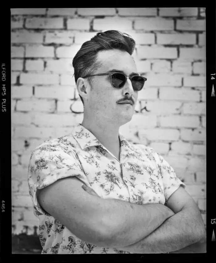 A man stands against a brick wall wearing sunglasses and folding his arms.