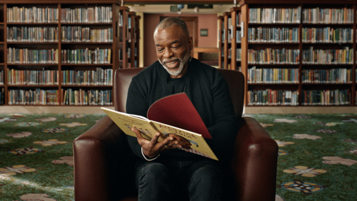 LeVar Burton sits in a chair with an open children's book in his hands