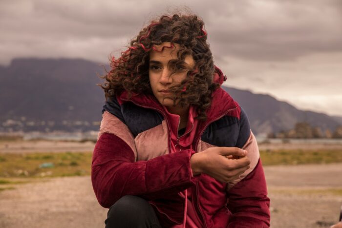 Khadija Jaafari as Jamila, sitting in a field, her hair blown by the wind in Californie.
