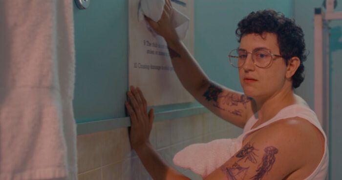 Emilie Modaff, a nonbinary actor, in a 1980's bathhouse, wiping a sign clean.