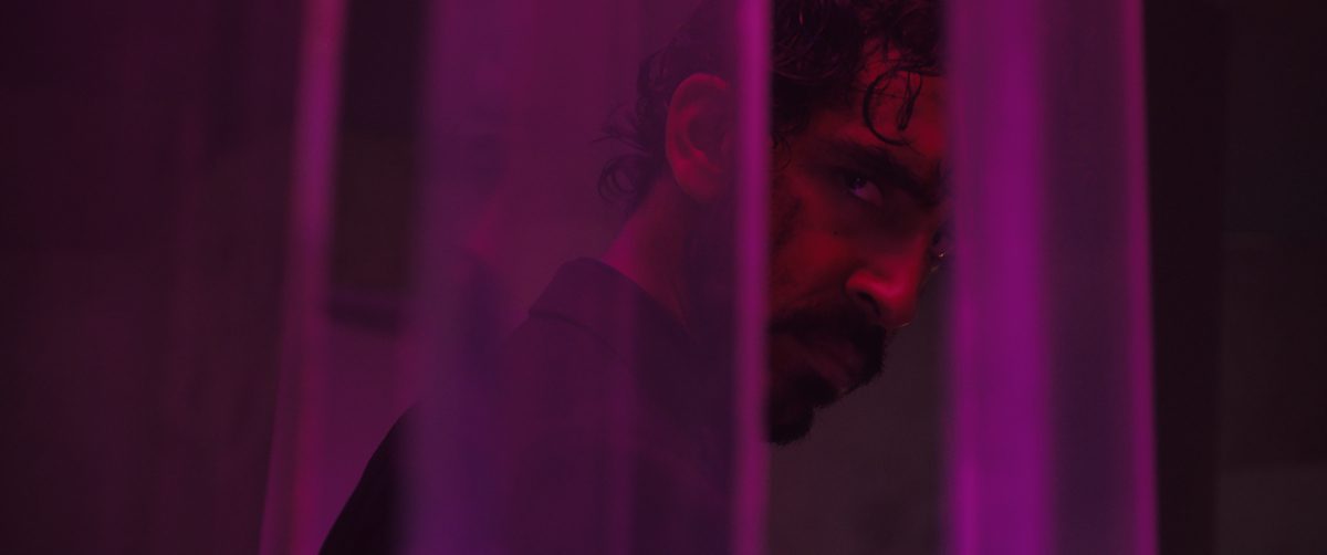 Dev Patel as the Kid in MONKEY MAN, directed by Dev Patel. © Universal Studios. All Rights Reserved. Sweaty and bloody from a fight, the Kid stares through sheer purple curtains.