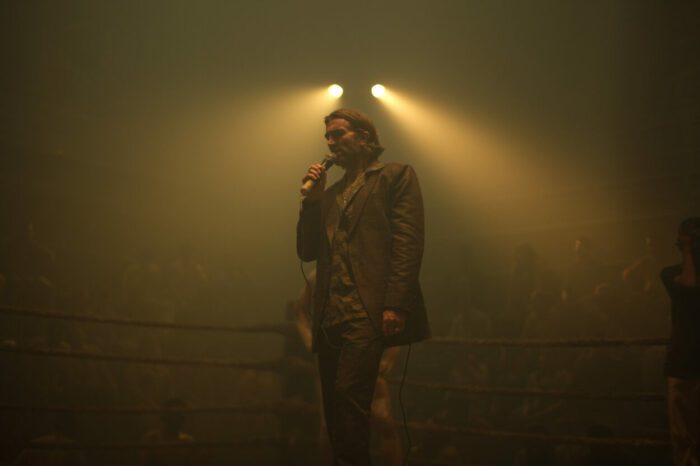 Sleazy promotor of an underground fight ring stands in the ring wearing a suit and too much gold jewelry, while announcing the next fight.