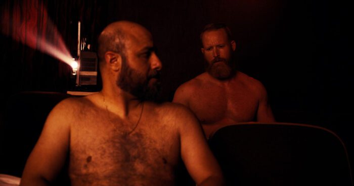 Ahmad (Aden Hakimi) and a man (Mike Geraghty) both sitting shirtless in a theater in Desire Lines. The man sitting behind Ahmad is looking directly at him.