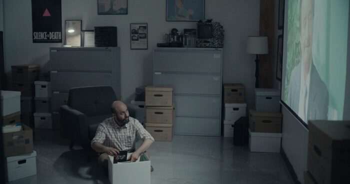 Ahmad (Aden Hakimi) in Desire Lines, sorting through a box on the floor while watching Lou Sullivan give an interview on a projector.