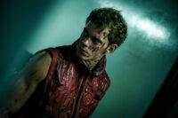 Bill Skarsgård as Boy in Boy Kills World (2024). Courtesy of Roadside Attractions. Covered in blood and wearing a red sleeveless vest, Boy stands in concrete room waiting to strike.