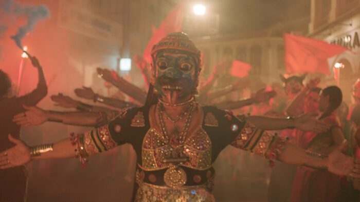 Street performers in traditional Indian garb create a Hindu multi-arm deity while dancing in a parade in Monkey Man.