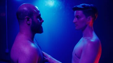 The characters Kieran (Theo Germaine) and Ahmad (Aden Hakimi ) in Desire lines, both shirtless in multicolored lighting. Kieran's top surgery scars are barely visible.