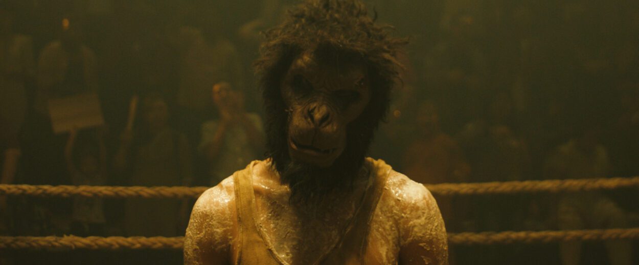 Dev Patel as the Kid in MONKEY MAN, directed by Dev Patel. © Universal Studios. All Rights Reserved. The Kid wears a monkey mask as he stands in a ring waiting for a fight.