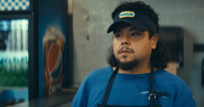 Lenny (Saul Trujillo), wearing his SubClub employee hat and apron, takes Roger's order.