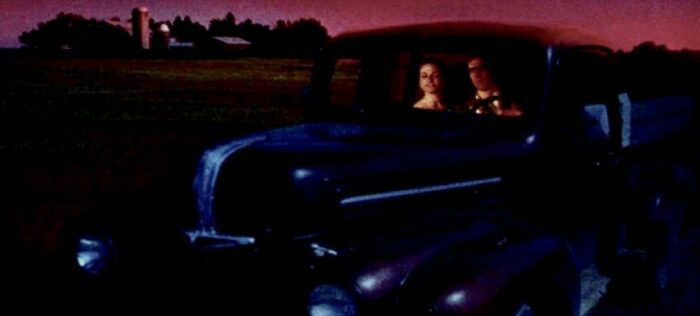 A couple sits in a 1950s vehicle with a purple sky in the background.