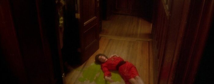 A dead alien, disguised as a human, laying on a wooden floor with green blood leaking from under her.