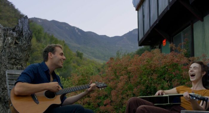 Two people play guitars together outside a cabin. 