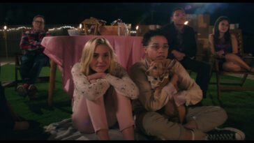 Two teens sit on the grass while friends and family watch an outdoor movie in The Moon & Back.