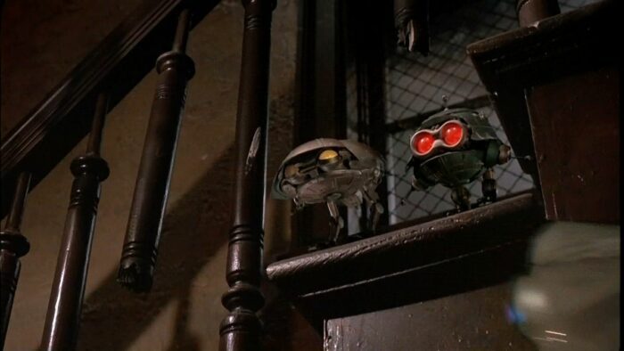 Two small robotic spaceships look through a banister with their eyes illuminated.