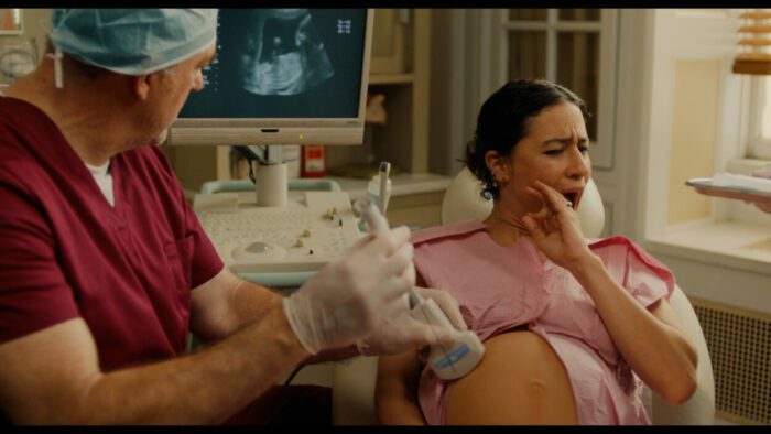 Ilana Glazer and John Carroll Lynch as Eden and Dr. Morris in BABES Photo Courtesy of NEON. Eden comedically reacts to the large needle Dr. Morris intends to use for amniocentesis.