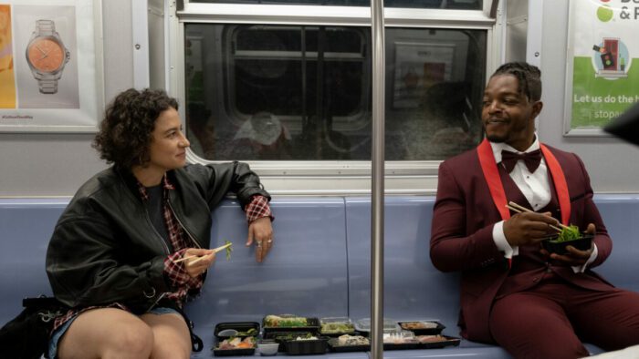 Ilana Glazer and Stephan James as Eden and Claude in BABES. Photo Credit to Gwen Capistran, Courtesy of NEON. Eden and Claude, who is wearing a maroon tux, riding the subway eating sushi together.