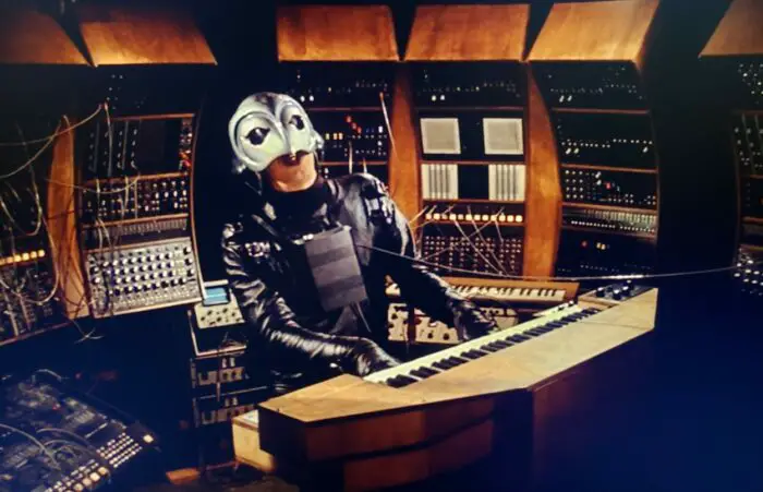 The Phantom at a bank of synthesizers playing a tune on a keyboard.