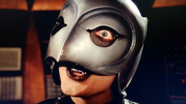 William Finley as the Phantom in Phantom of the Paradise (1974). Screen capture off of Amazon. © 20th Century Fox. The Phantom in black leather and steely bird mask with one visible eye bulging insanely.