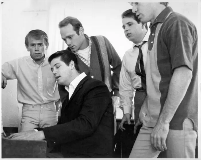 1964: Al Jardine, Brian Wilson, Mike Love, Carl Wilson and Dennis Wilson of the rock and roll band "The Beach Boys" sing around a piano . (Photo by Michael Ochs Archives/Getty Images). Blak and white image of the Beach Boys gathered around a piano rehearsing.