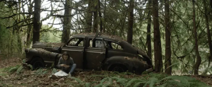 The young girl sits against an old car in the forest