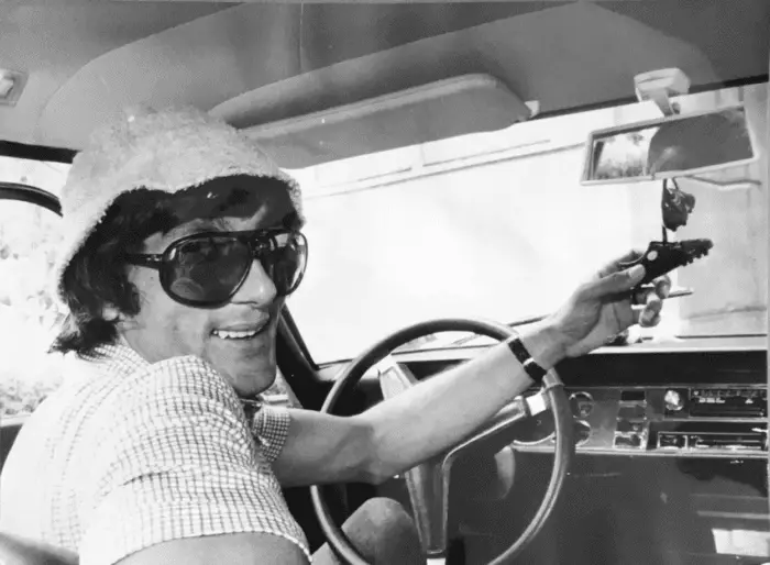 Wearing sunglasses and a bucket hat, Ilie Nastase looks at the camera from the driver's seat of a car. 