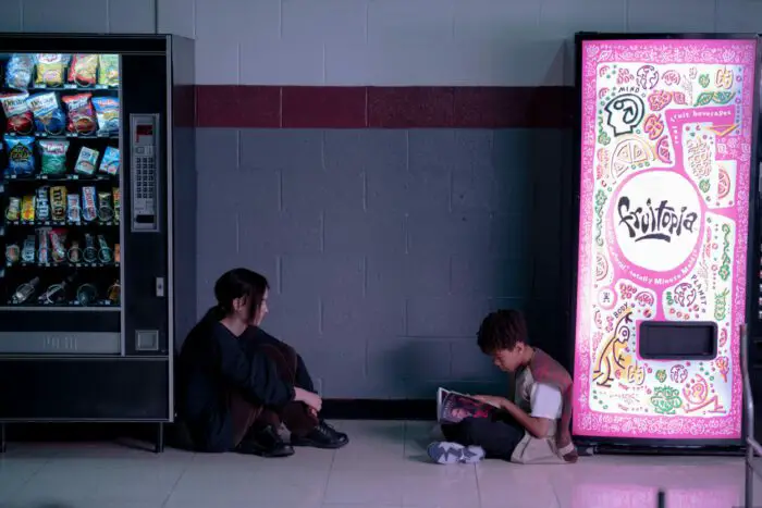 Maddy and Owen sit in between two vending machines. The one beside Maddy is bleak and dark and the one beside Owen is glowing neon pink.