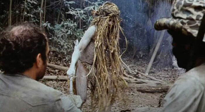 Robert Kerman (Prof. Monroe) and Salvatore Basil (Chaco) with uncredited performer as a Yąnomamö shaman in Cannibal Holocaust (1980). Screenshot off Peacock. Clad in gras and fronds a shaman covered in ash making his skin grey, performs a ritual before Chaco and Prof. Monroe.