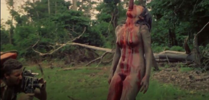 Luca Barbareschi as Mark Tomaso filming the impaled woman (actor uncredited) in Cannibal Holocaust. Screenshot off Peacock streaming. A naked woman on a river shore is impaled vertically, the pole sticking out her bloody mouth, while Mark videotapes the gruesome display with a 16mm camera.