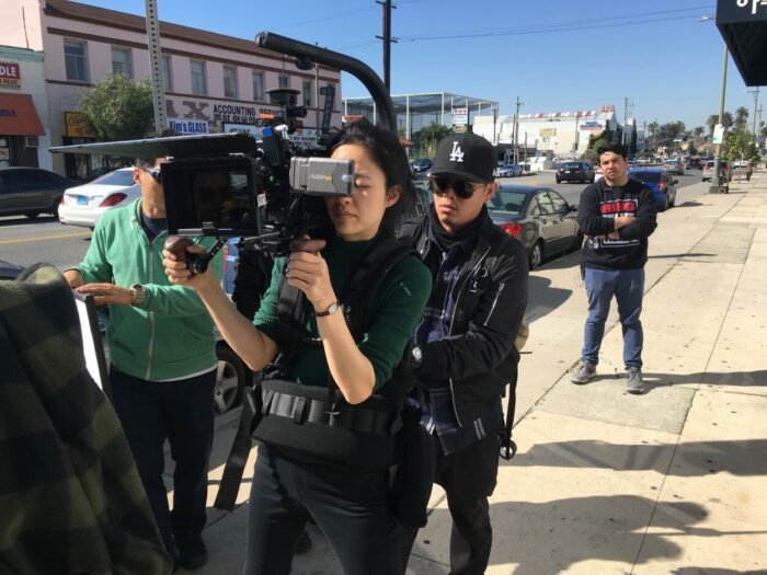 A woman carries a camera on the street a a crew follows.