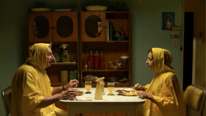 Norberto and Rita sit at the kitchen tables in rain ponchos in Chronicles of a Wandering Saint