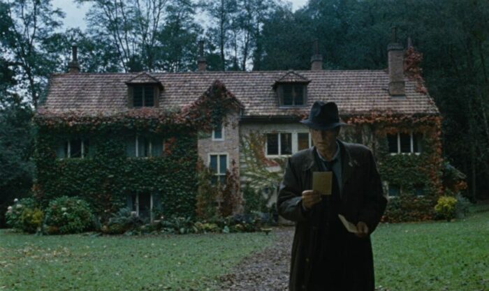 A man in a trench coat and a hat standing in front of a house looking at a photo.
