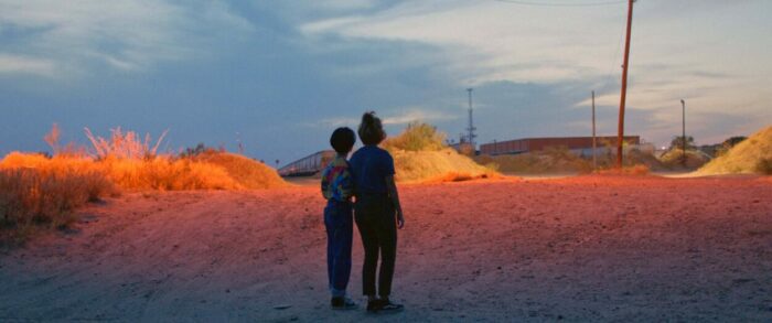 Caption: Estefanía “Beba” Contreras (left) and Silvia Del Carmen Castaños (right) watch a train in the distance near the U.S.-Mexico border at dusk in a scene from Hummingbirds
