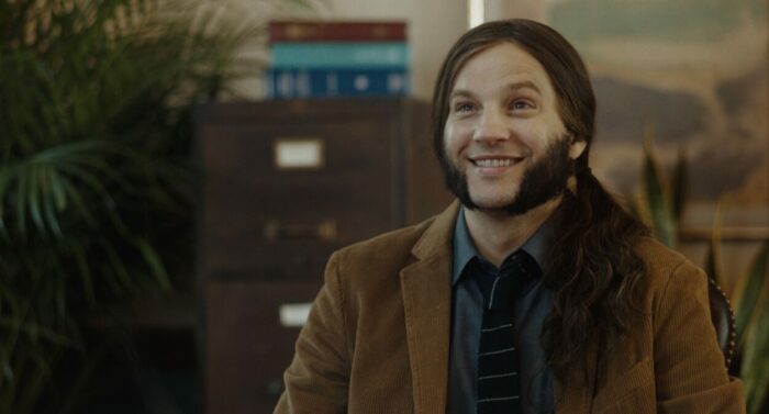 Logan Marshall-Green as Ted in Reverse the Curse (2023). Courtesy of Vertical and R&CPMK. A long-haired man with mutton chops sits in a suit and tie awaiting input from a literary agent about his failed book.