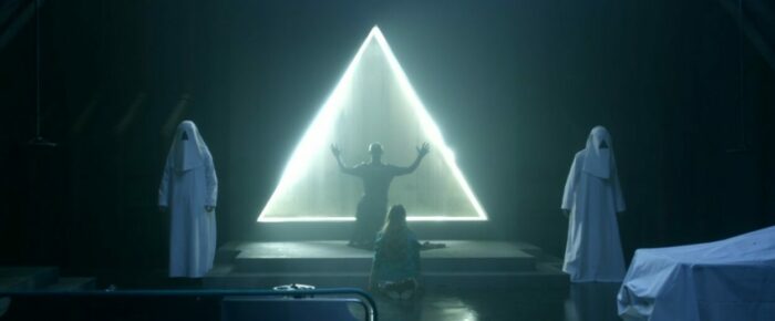 A mysterious figure stands before a large triangle that is brightly lit at the edges.