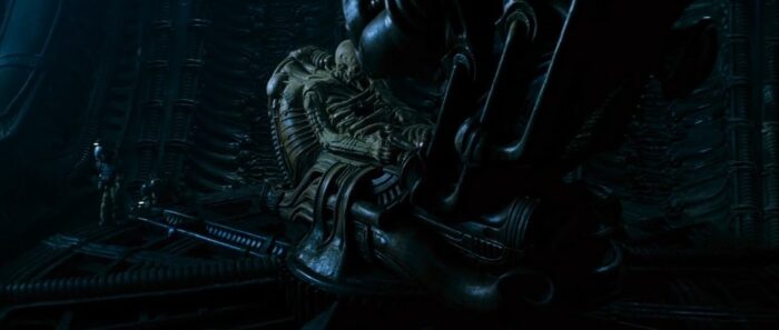 Image from ALIEN showing the large corpse of a mysterious alien figure. to the left is a human in a spacesuit. 