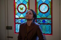 Julia Louis-Dreyfus as Zora in Tuesday. Credit: By Kevin Baker. Courtesy of A24. Depressed mother Zora stands leaning back against her stained-glass front door, the glass features colorful images of birds.