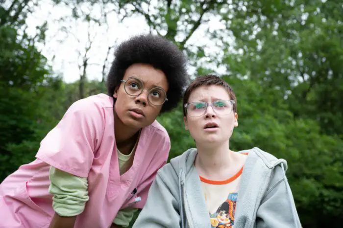 (L-R) Leah Harvey, Lola Petticrew as Nurse Billie and Tuesday in Tuesday. Credit: By Kevin Baker. Courtesy of A24. Nurse Billie in pink scrubs stands beside bespectacled teenager Tuesday in a wooded field.