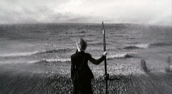 A woman stands at the edge of the sea with a harpoon.