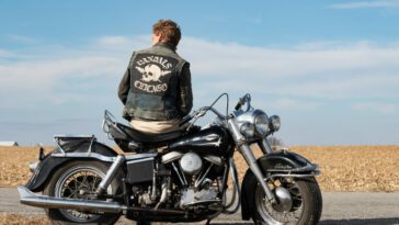A man with a jacket sits on a parked motorcycle in the open country in The Bikeriders.