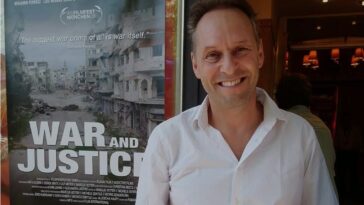 Marcus Vetter smiles in front of a poster for War and Justice.