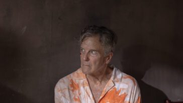 Bloodied Nicholas Hammond sits in a plain room