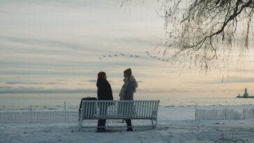 Two people stand outside near a park bench in Close to You.