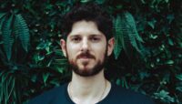 Gabriel Mayo, depicted in front of dense foliage wearing a black t-shirt and gold chain.